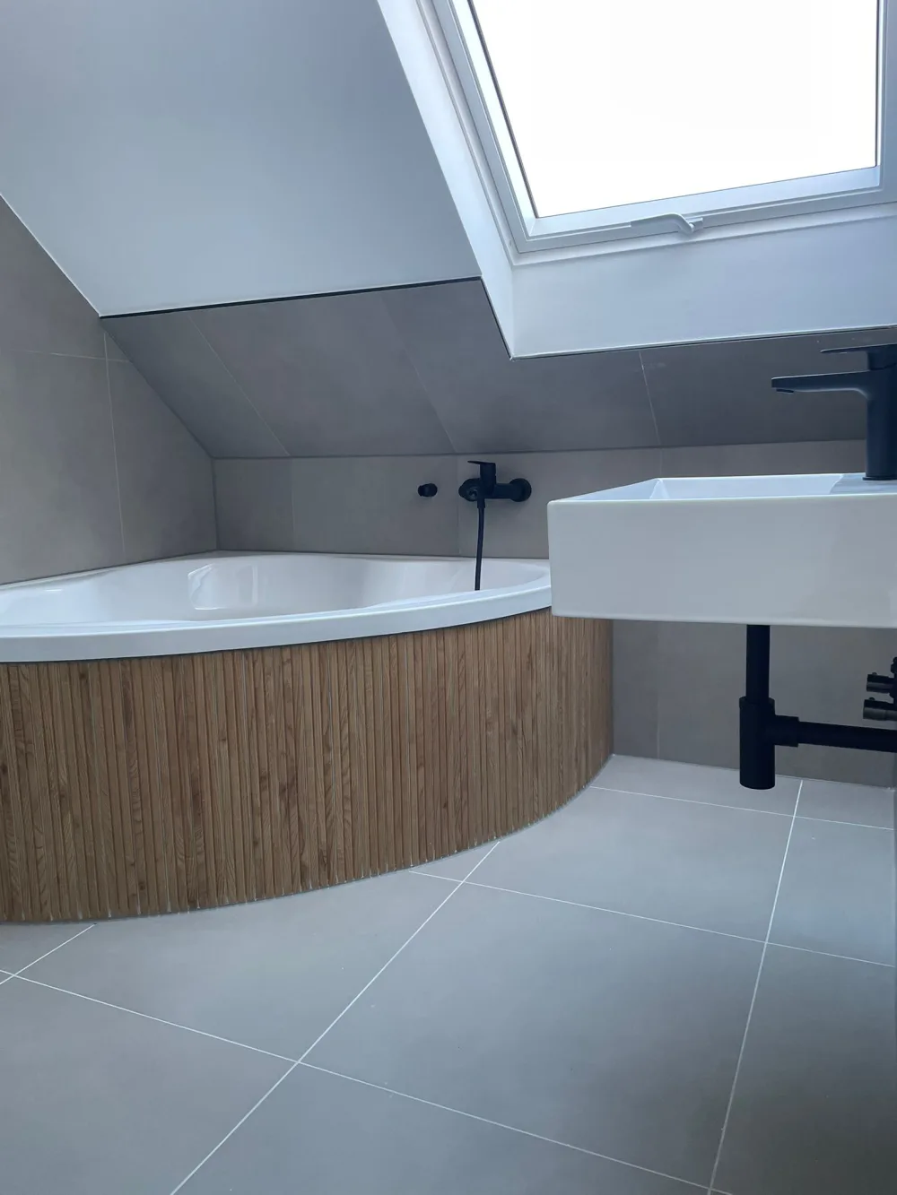 Modernisiertes Dachgeschossbad mit Badewanne, Dachfenster, grauen Fliesen, Waschtisch und Holzverkleidung