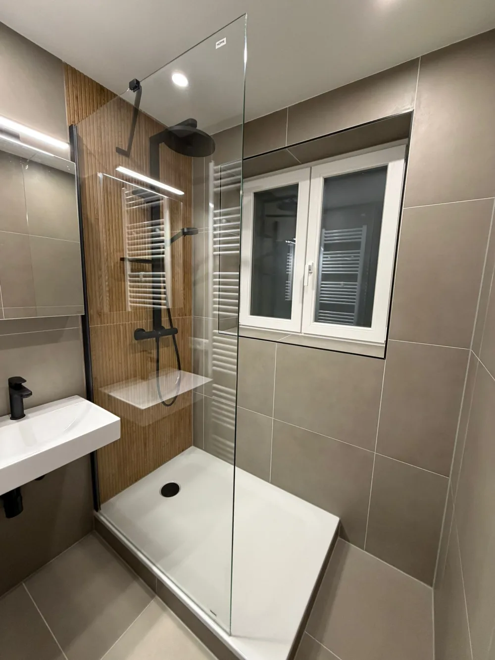 Modernisiertes Bad mit Glasdusche, grauen Fliesen, Holzoptik-Wand, schwarzer Armatur, Waschtisch und Fenster