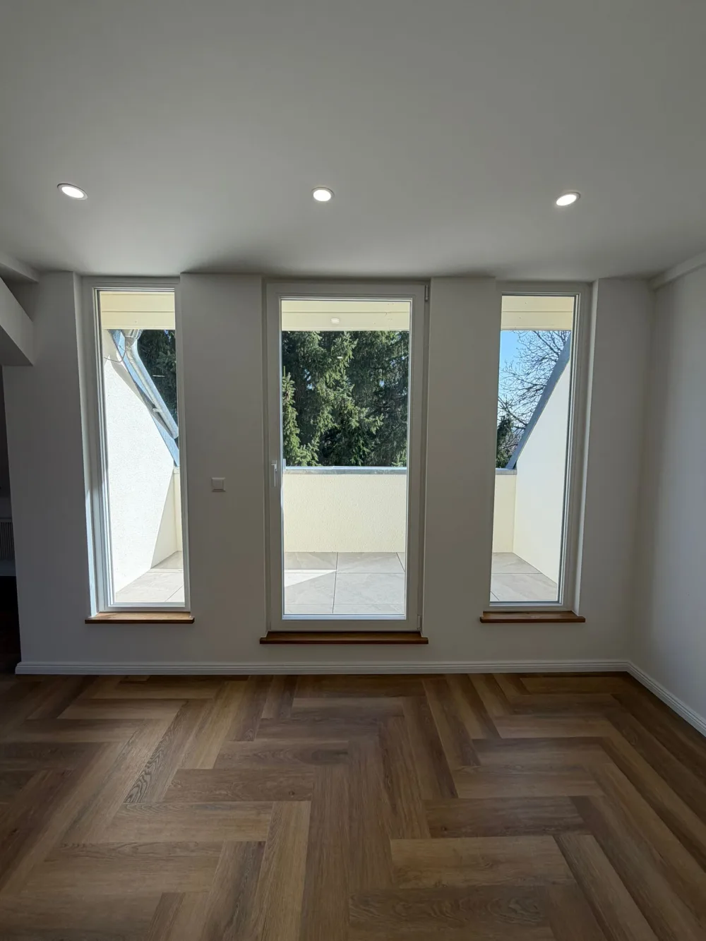 Modernisierter Dachgeschoss-Wohnbereich mit großer Fensterfront, Balkonzugang, Fischgrätboden, weißen Wänden und Deckenspots
