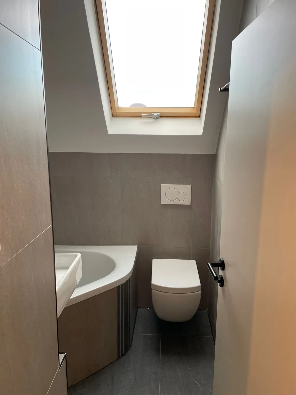 Modernisiertes Gäste-WC mit grauen Fliesen, wandhängendem WC, Waschbecken, Dachfenster und sauber ausgeführten Wandflächen